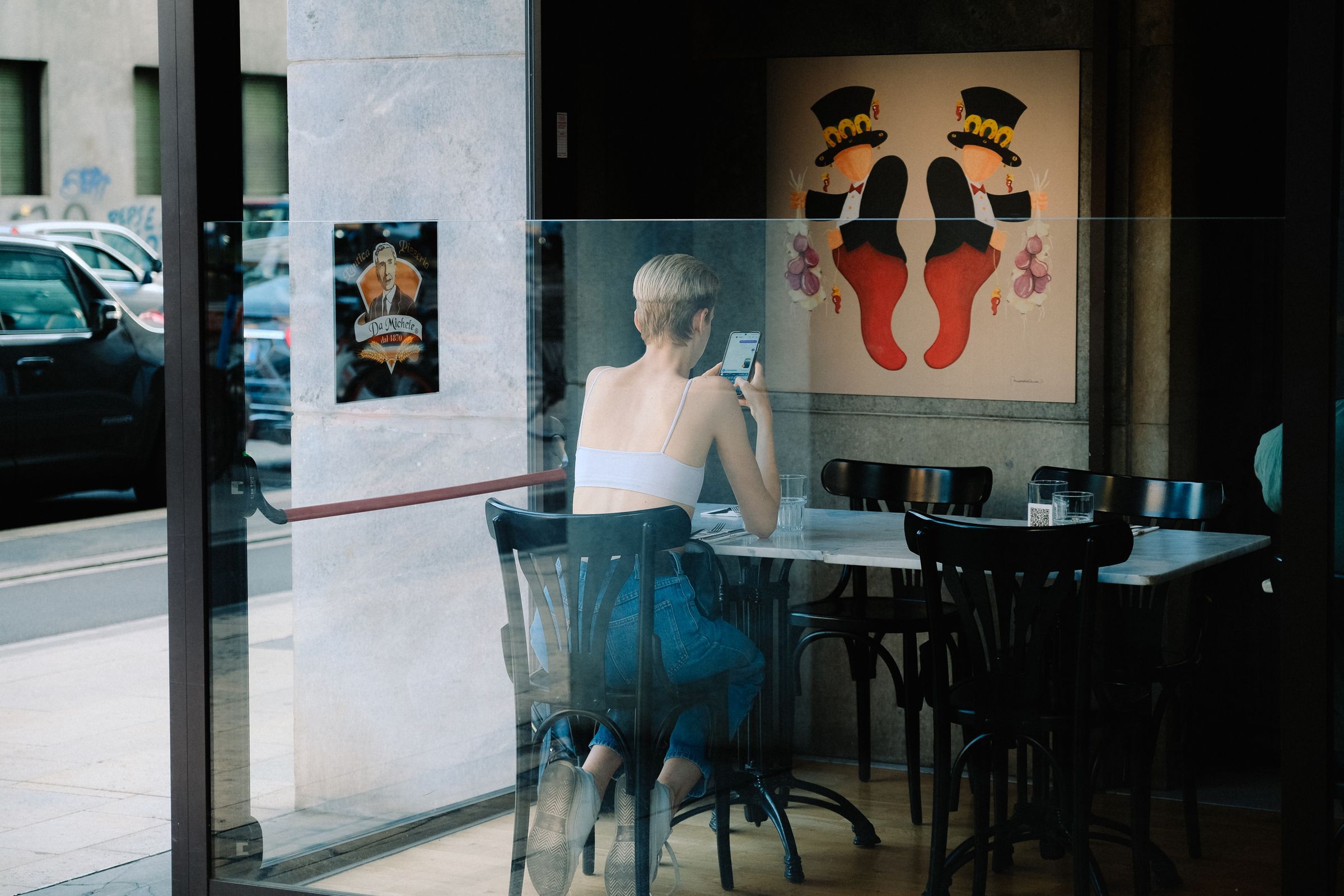 A woman uses her smartphone at a cafe table, viewed through a window
