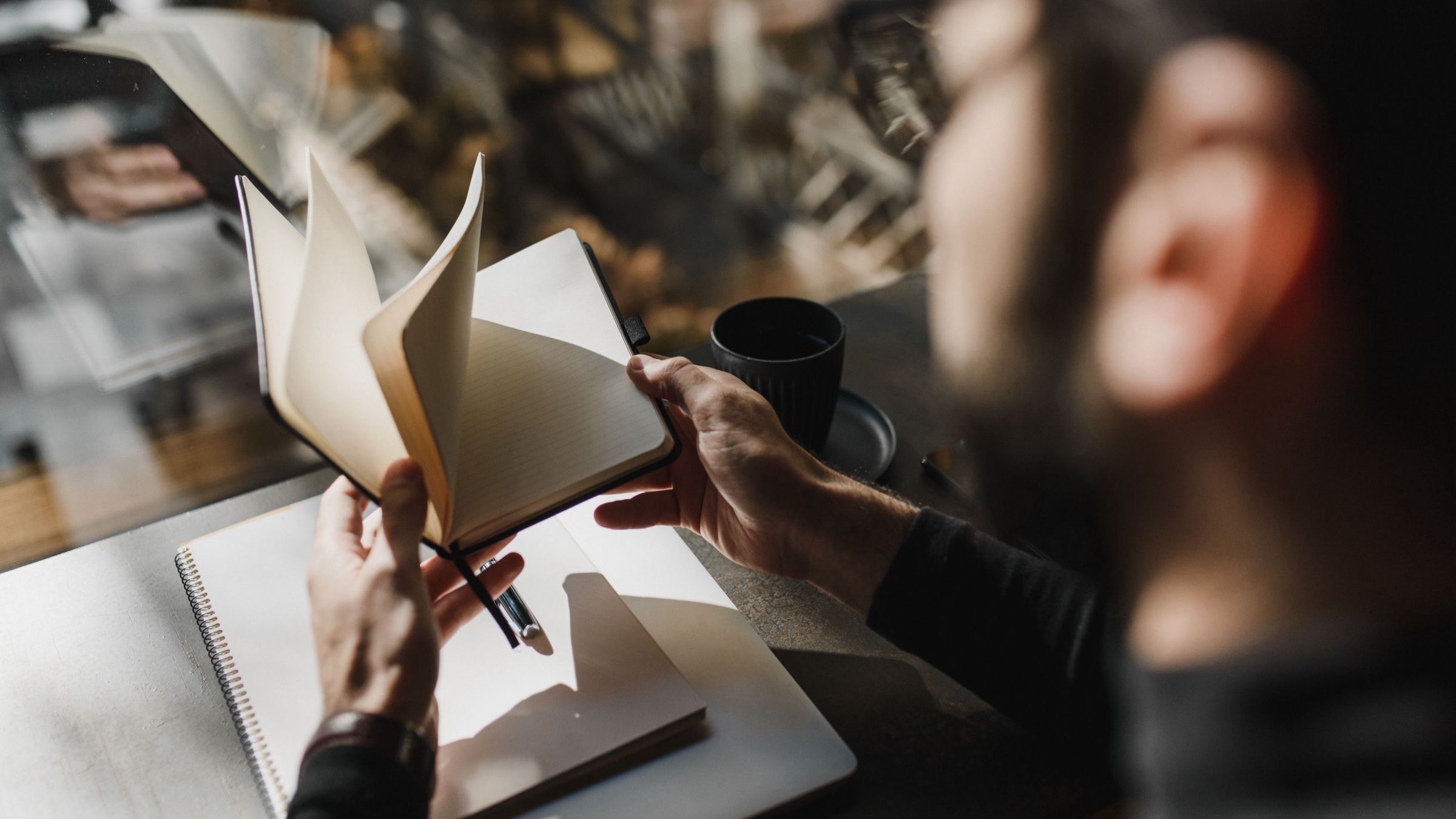 An adult reviewing a blank notebook at a sunlit cafe table